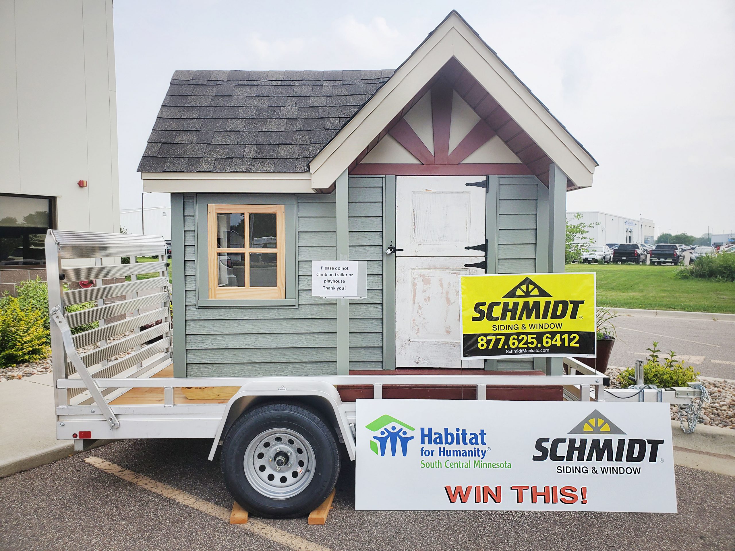 The image shows a small playhouse atop a low-sided trailer. The logo of HFHSCMN and Schmidt Siding & Window is on a sign with the words "Win this!"